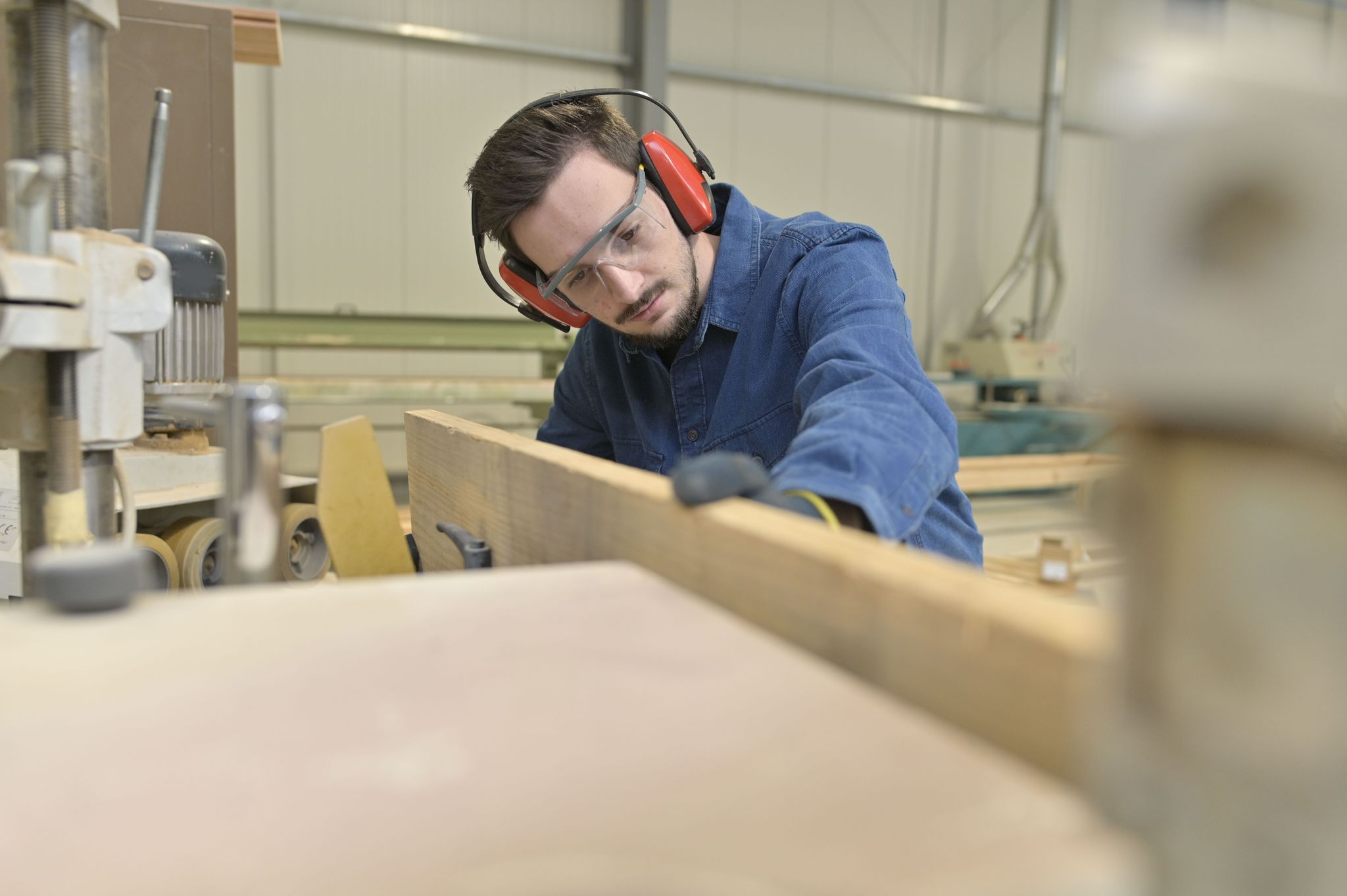 A woodworker aligning a piece of plywood against a saw while wearing hearing protective ear muffs.