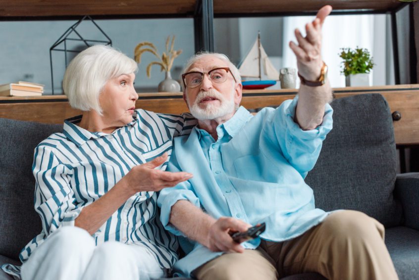Selective focus of senior man gesturing while watching tv with wife on couch in living room