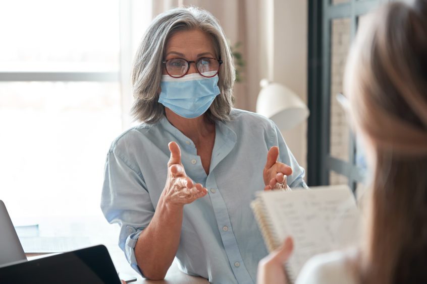 Old female mentor wearing face mask training young interns at office meeting.