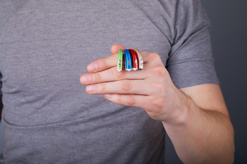 Man in grey shirt holding 4 hearing aids of different colors.