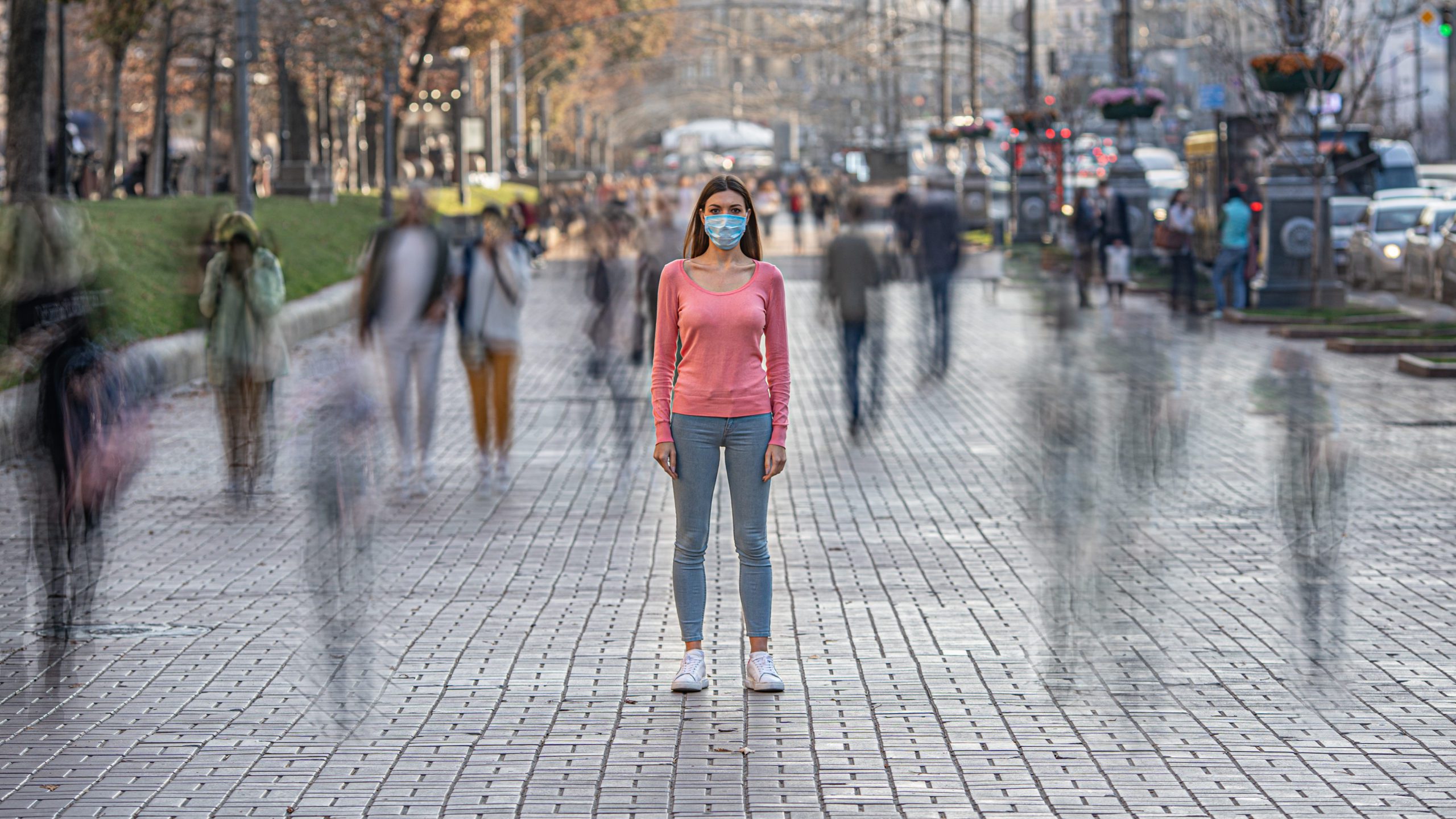 Woman in a pink shirt and N95 mask standing still on a busy walkway, with blurred, time-lapsed silhouettes of people moving around her.