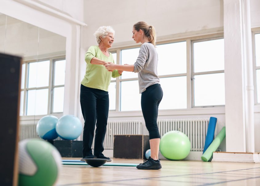A fitness instructor assisting a senior student use a balance board.