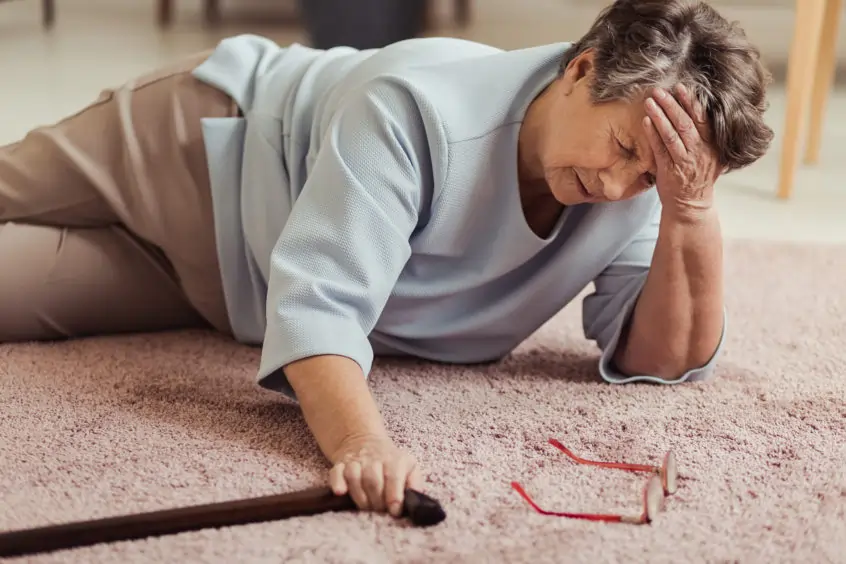 Senior woman holding her head in her hands after a fall, holding a walking cane, with red glasses on the ground next to her.
