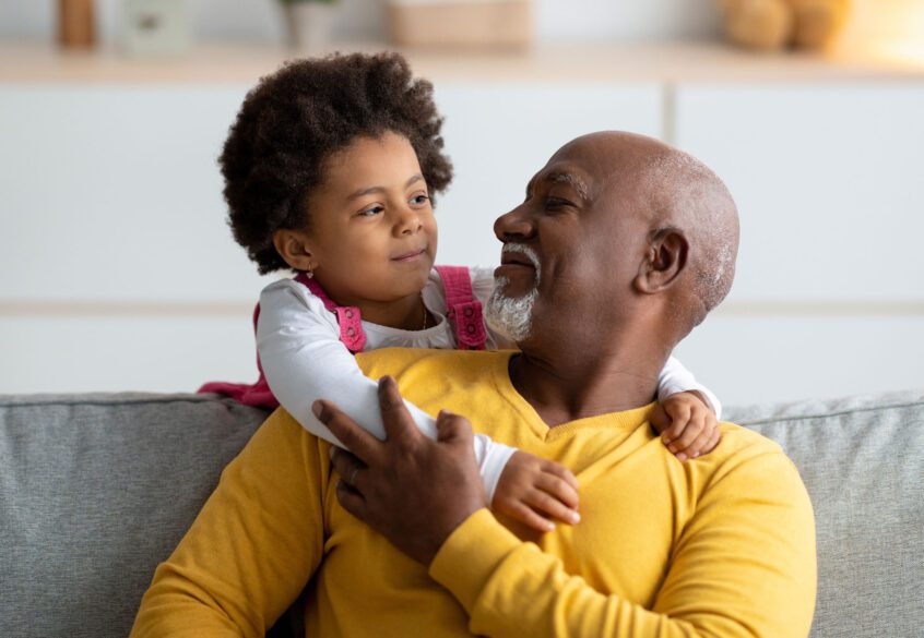 A grandfather and granddaughter embracing over a couch.