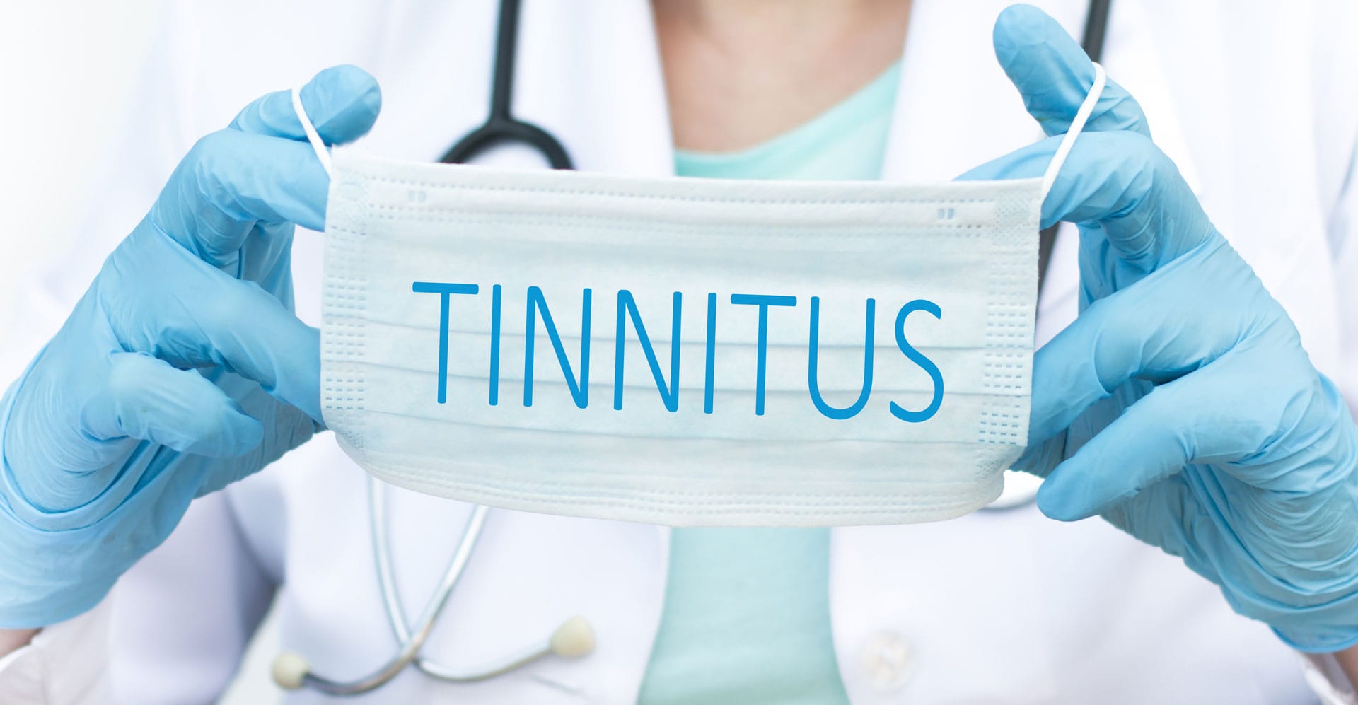 The image depicts a medical professional, likely a doctor or nurse, holding a surgical face mask with the word 'TINNITUS' prominently displayed across the front in bright blue capital letters. The healthcare worker is wearing light blue surgical gloves and a stethoscope is visible around their neck, partially obscured by their torso. They are dressed in a light green medical scrub top, suggesting a clinical setting. The background is softly blurred, appearing to be a light-colored wall or room, keeping the focus squarely on the mask and the message it conveys. The lighting is bright and even, highlighting the details of the gloves, the mask’s texture, and the lettering. The composition is centered, with the mask held directly in front of the person, creating a direct visual connection between the medical professional and the condition of tinnitus. The overall impression is one of medical awareness and concern regarding tinnitus, potentially indicating a focus on diagnosis, treatment, or research related to this hearing condition. The use of a face mask as the medium for the message is particularly striking, possibly alluding to the impact of health concerns, like tinnitus, in a world increasingly aware of medical issues.